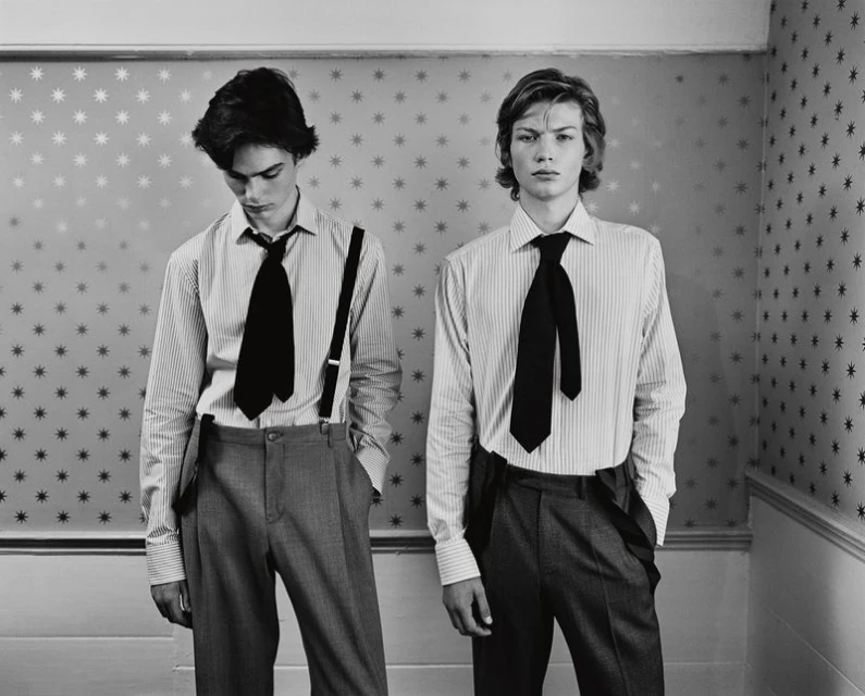Two young adult men in a photo studio wearing ties and suspenders, posing against a star-patterned wall, creating a vintage fashion look.