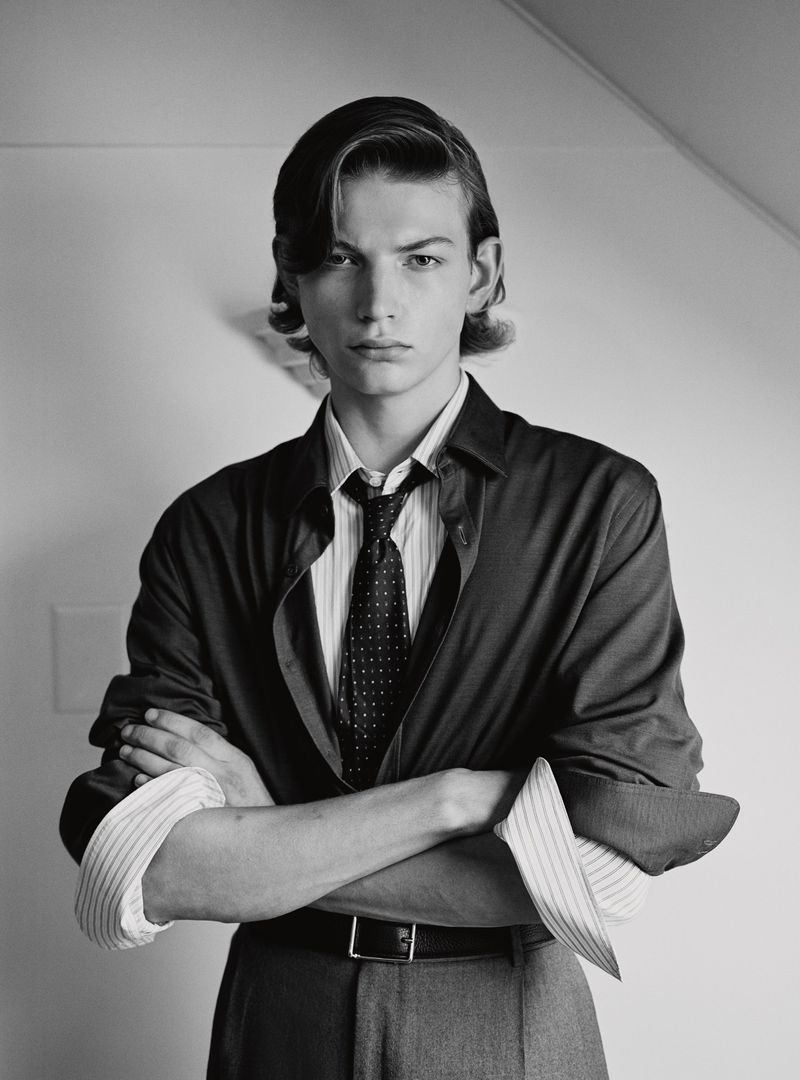 Black-and-white portrait of a young man in a dress shirt, tie, and suit jacket posing indoors with his arms crossed.