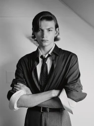 Monochrome portrait of a young male model with arms crossed wearing a dark buttoned shirt and tie against a plain light wall