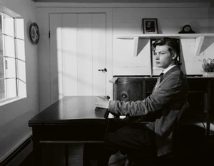 Young man in a jacket seated at a dark wooden desk in a room lit by natural window light