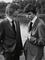 Two men in tailored wool suits and collared shirts with ties standing outdoors beside a body of water in black and white.
