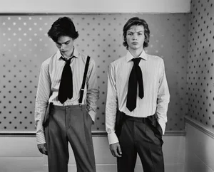 Two young male models in striped shirts and ties standing against a patterned star-print wall for AnOther Man Magazine.