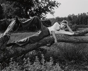 Young male model lounging on a fallen tree trunk in a grassy field wearing a white shirt and trousers.