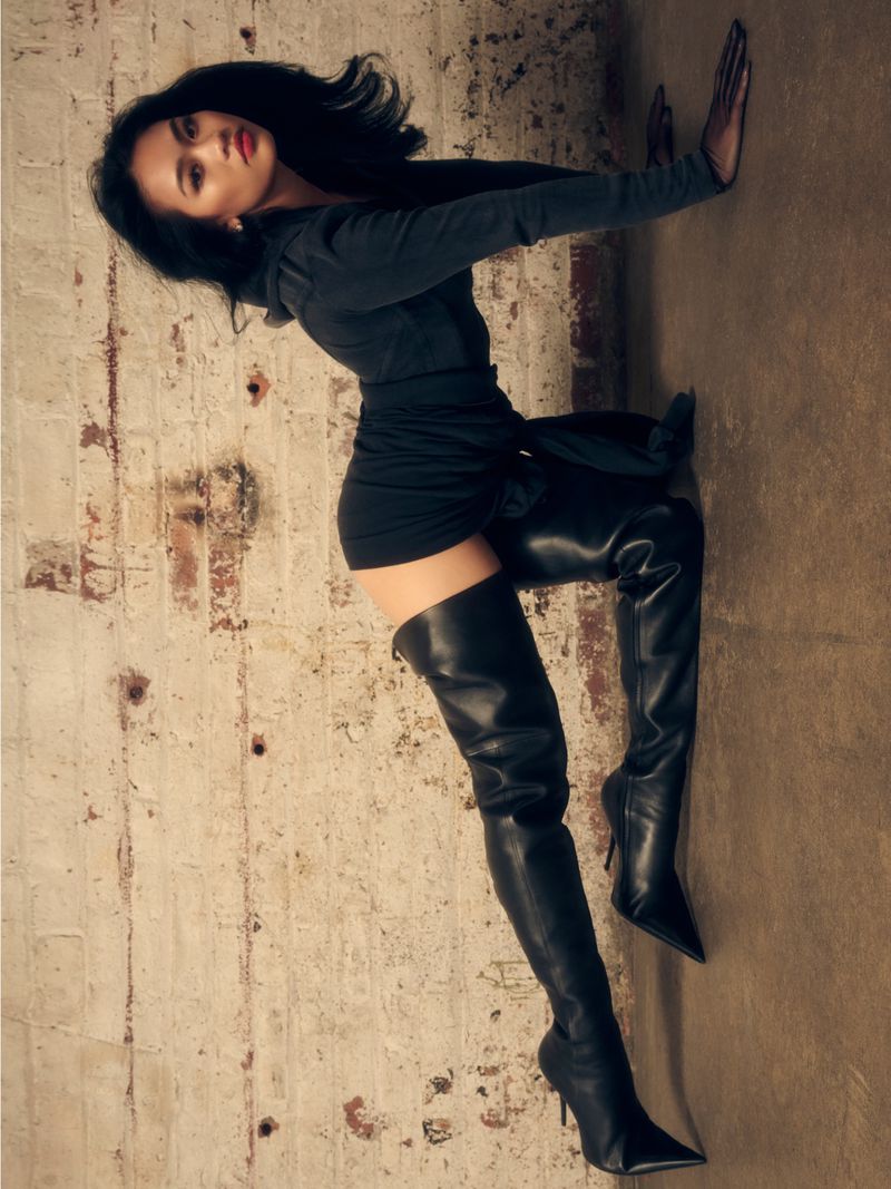 A woman in a short black dress and thigh-high black boots leans against a textured wall in a fashion-forward pose, photographed in a studio setting.