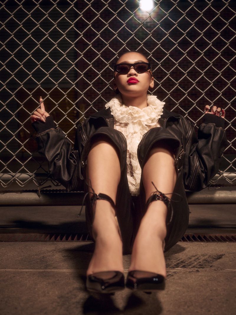Eye-level shot of a woman in a black bomber jacket, lace top, sunglasses, and heels sitting in front of a chain-link fence.