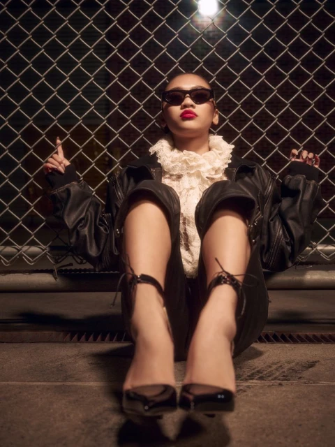 Eye-level shot of a woman in a black bomber jacket, lace top, sunglasses, and heels sitting in front of a chain-link fence.