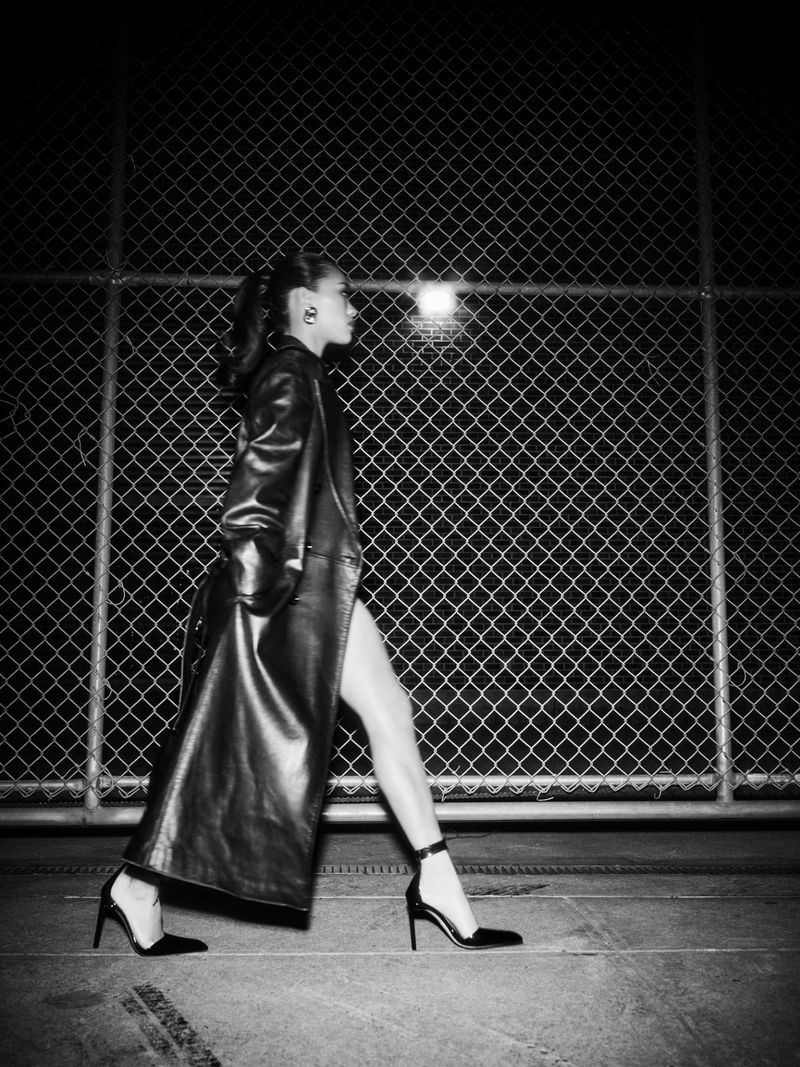 Black and white fashion shot of woman in long leather coat walking in high heels against chainlink fence. Dark, moody lighting.