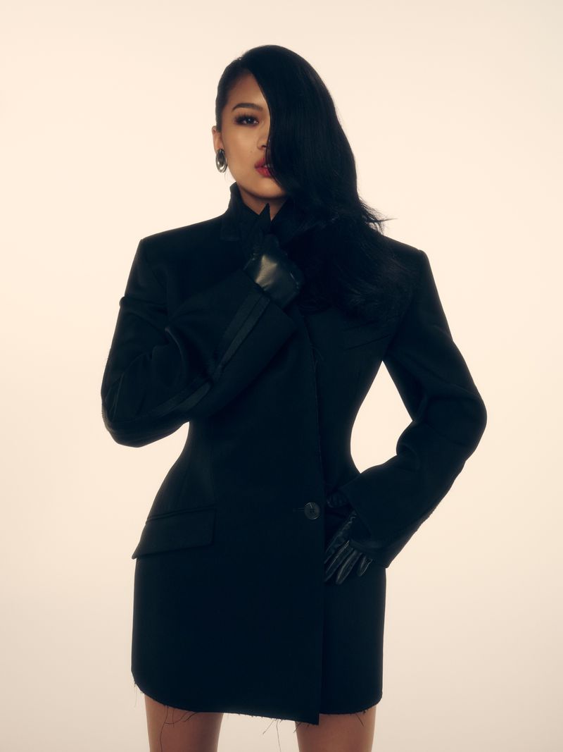 Portrait of an adult female with long black hair, wearing a black blazer dress and gloves, posing in front of a neutral background.