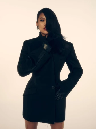 Portrait of an adult female with long black hair, wearing a black blazer dress and gloves, posing in front of a neutral background.