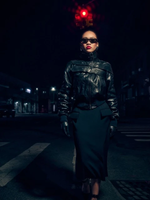 Woman in black leather jacket and skirt stands on a crosswalk at night with a red traffic light in the background.