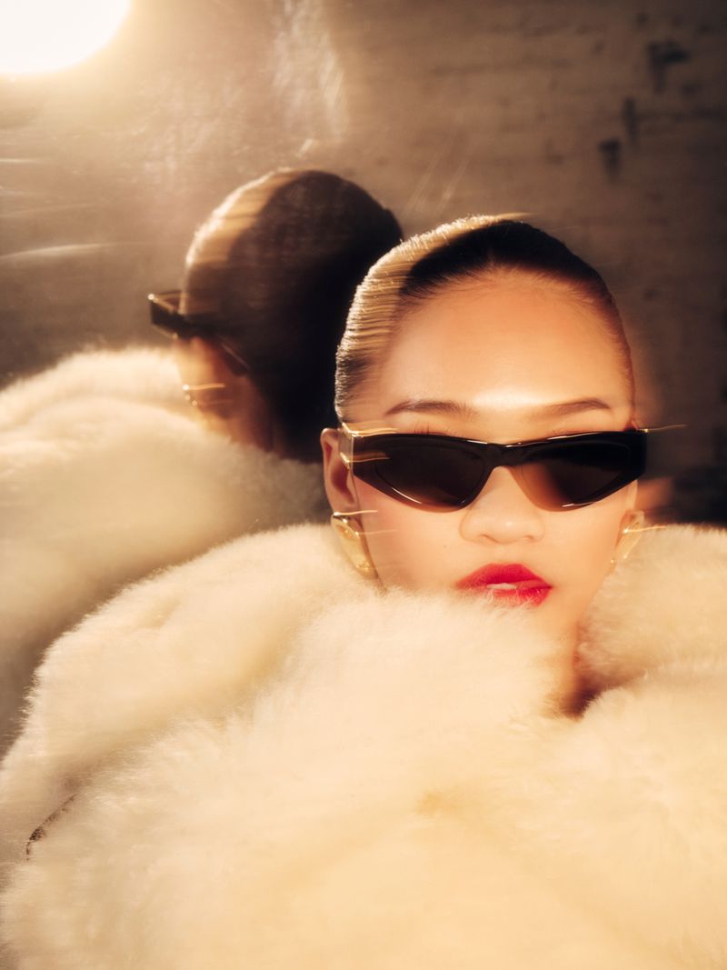 Portrait of a woman with sunglasses and red lipstick wearing a fur coat, partially reflected in a mirror with blurred background.