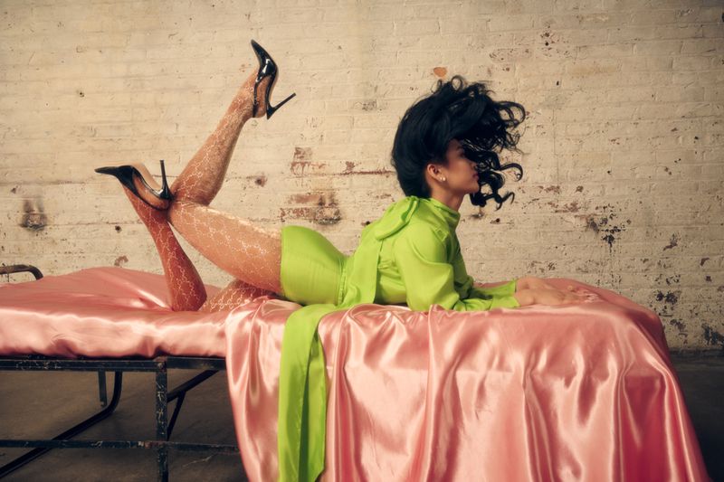 Woman in green dress and black heels poses on a pink satin sheet in front of a white brick wall in a fashion-forward studio shot.