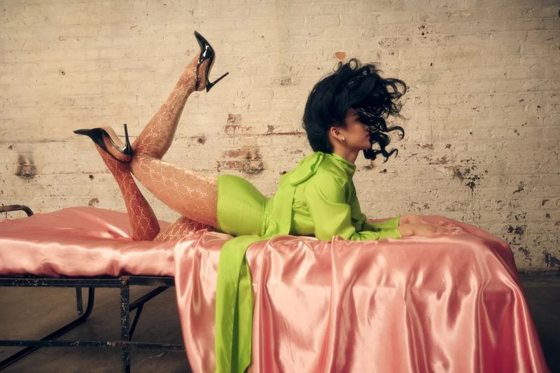 Woman in green dress and black heels poses on a pink satin sheet in front of a white brick wall in a fashion-forward studio shot.