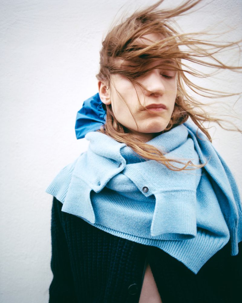 Portrait of a woman with windswept hair wearing a blue scarf, standing against a neutral wall, captured with shallow depth of field.