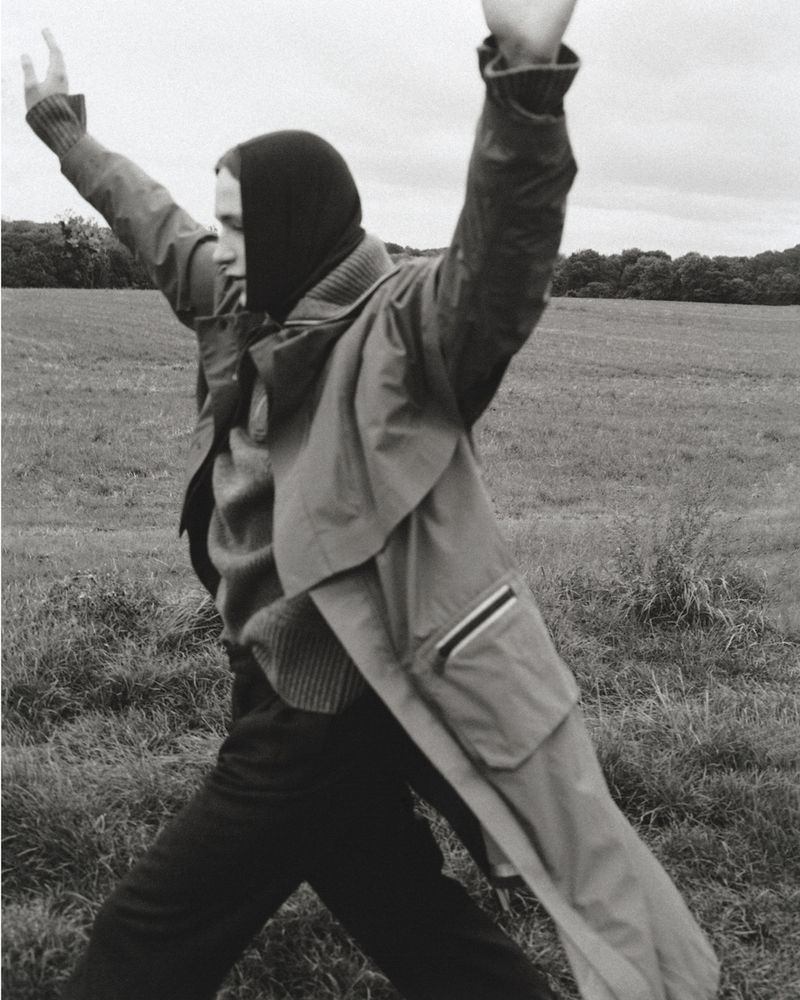 Monochrome fashion editorial image of a person wearing a coat and dark head covering with arms raised in an open field.