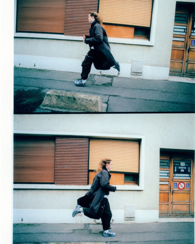 Woman in a long dark coat and athletic shoes runs along an urban sidewalk near a building with brown window shutters and door.