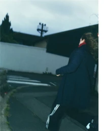 Woman in dark coat and track pants walking on an outdoor street under an overcast sky.