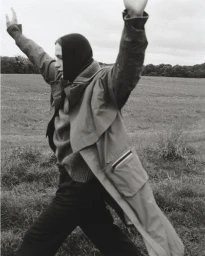 Monochrome fashion editorial image of a person wearing a coat and dark head covering with arms raised in an open field.