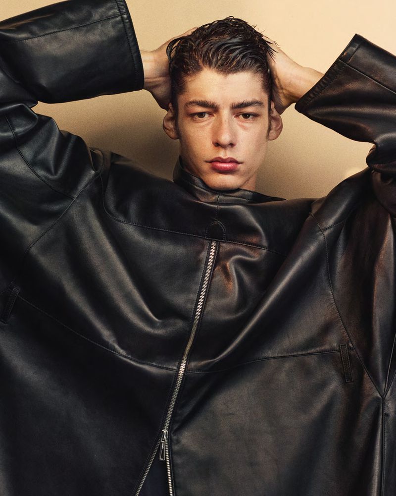 Portrait of a young adult male with dark hair, posing with hands behind his head, wearing a black leather jacket against a neutral backdrop.