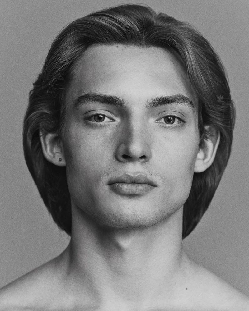 Monochrome portrait of a young adult man with long hair, facing forward with a neutral expression against a solid grey background.