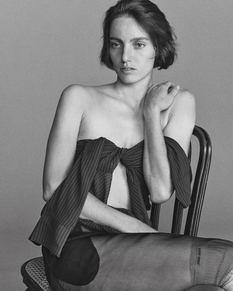 Black and white portrait of a woman with short dark hair seated on a cane chair wearing a pinstripe off-shoulder top.