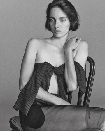 Black and white portrait of a woman with short dark hair seated on a cane chair wearing a pinstripe off-shoulder top.