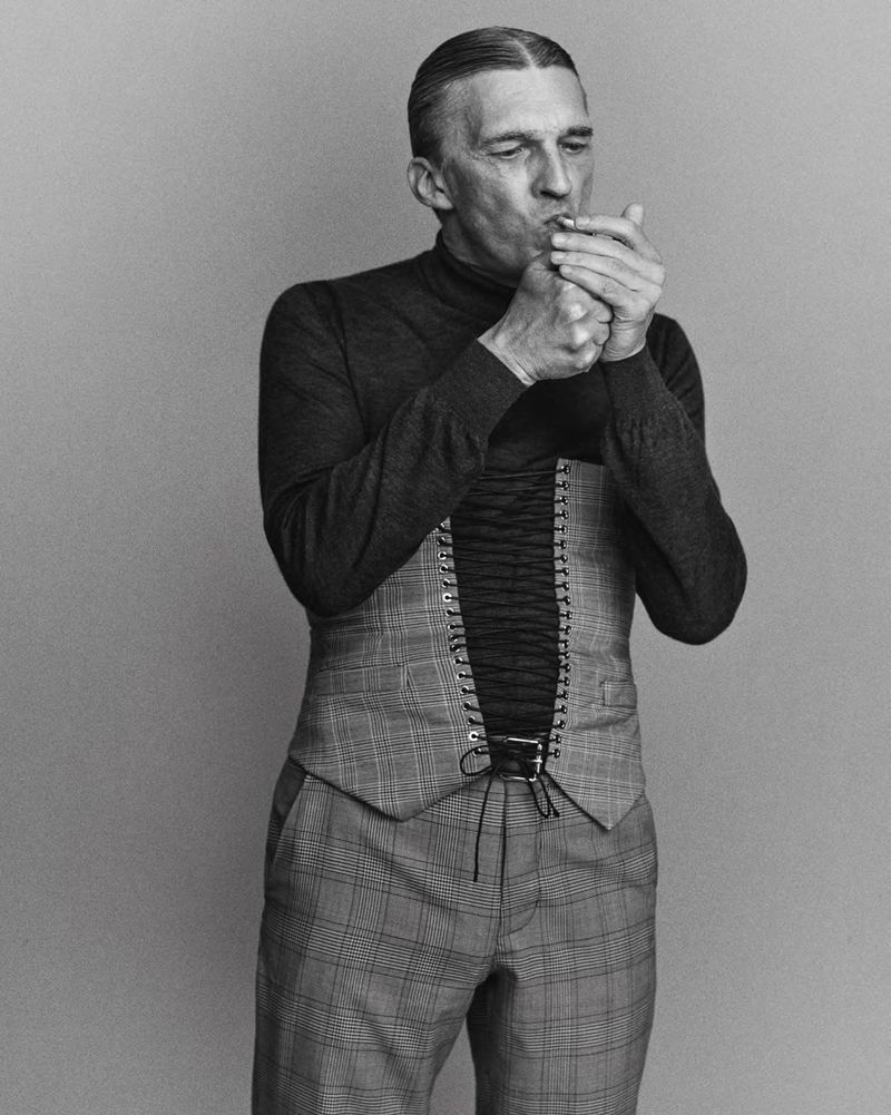 Monochrome portrait of a mature man smoking, wearing a vest and turtleneck, set against a plain backdrop, with a dramatic lighting.