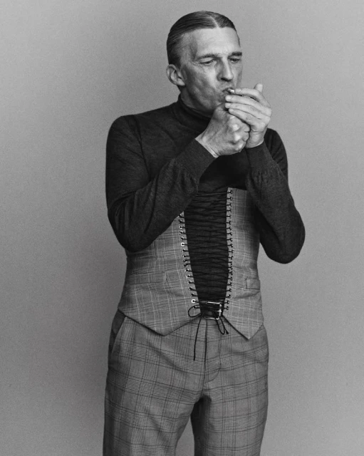 Monochrome portrait of a mature man smoking, wearing a vest and turtleneck, set against a plain backdrop, with a dramatic lighting.