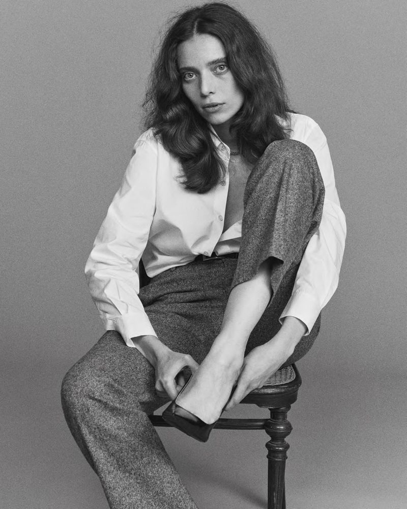 Black and white portrait of an adult woman with long wavy hair sitting on a stool in a white shirt and grey slacks, gazing forward.