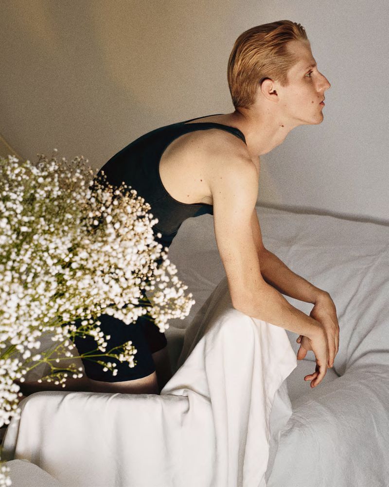 Young fair-skinned man in black top leans against a white couch indoors next to baby's breath flowers. Side profile.