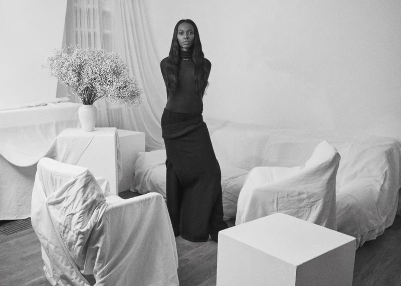 Elegant Black woman in a long skirt poses in a sparse indoor setting with draped furniture and a vase of white flowers, black-and-white.