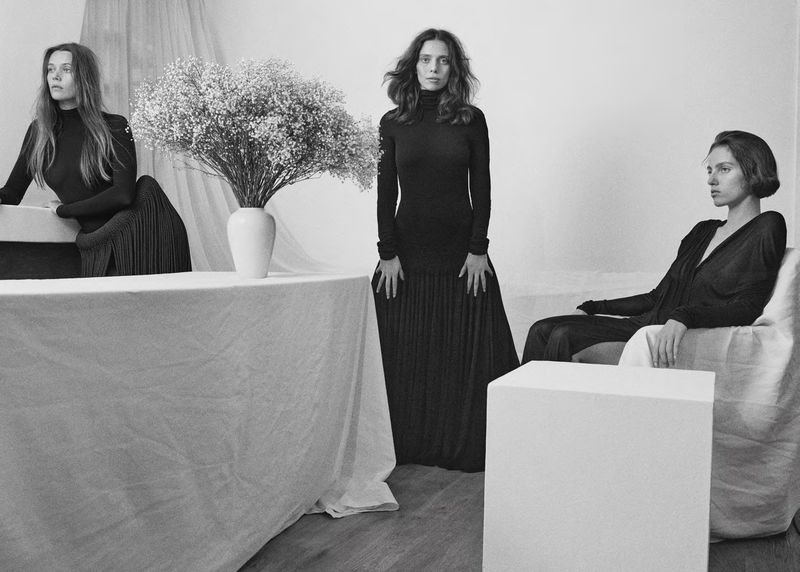 Monochrome studio shot featuring three female models in dark dresses, one standing, and the other two seated near a white table and cube.