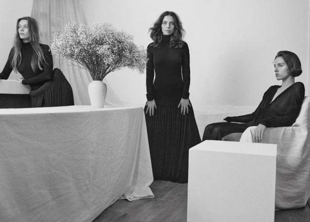 Monochrome studio shot featuring three female models in dark dresses, one standing, and the other two seated near a white table and cube.