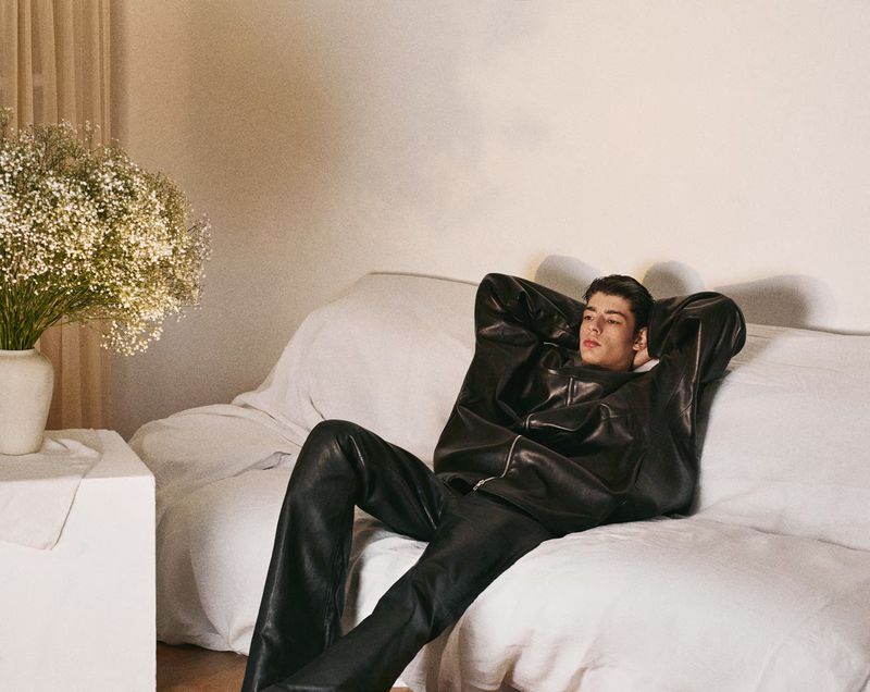 A relaxed adult male wearing a black leather jacket and pants rests on a white sofa next to a white side table with flowers.