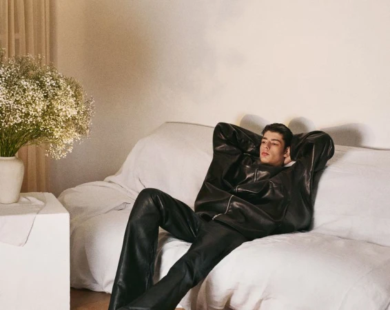 A relaxed adult male wearing a black leather jacket and pants rests on a white sofa next to a white side table with flowers.