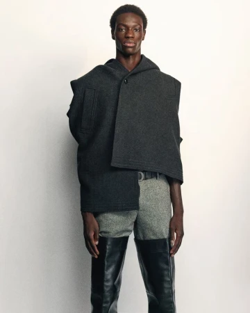Editorial fashion shot featuring a Black male model wearing a hooded gray woolen cape, light gray trousers, and black leather thigh-high boots.