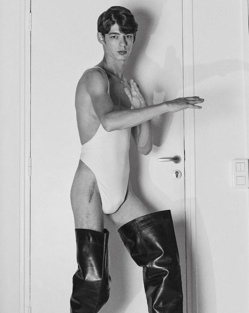 Black and white studio portrait of a slender adult man wearing a leotard and boots, posing near a white door with his hands up.