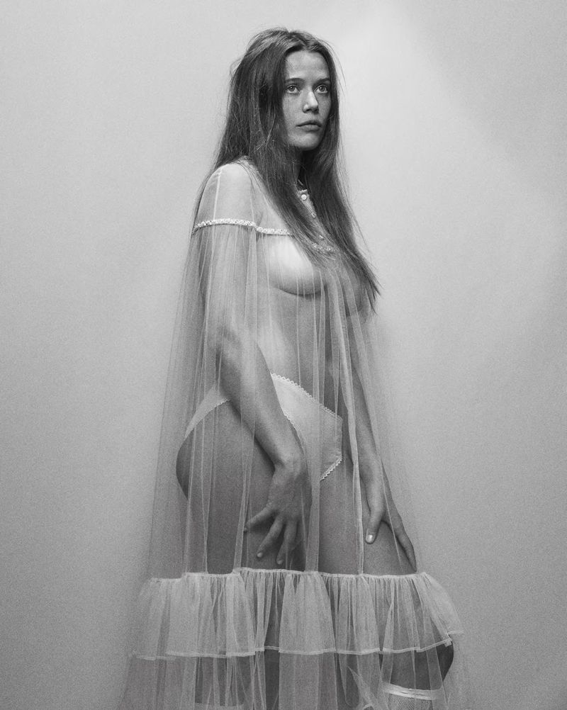 Monochrome portrait of a woman with long hair, partially nude, draped in sheer white fabric against a plain off-white backdrop.
