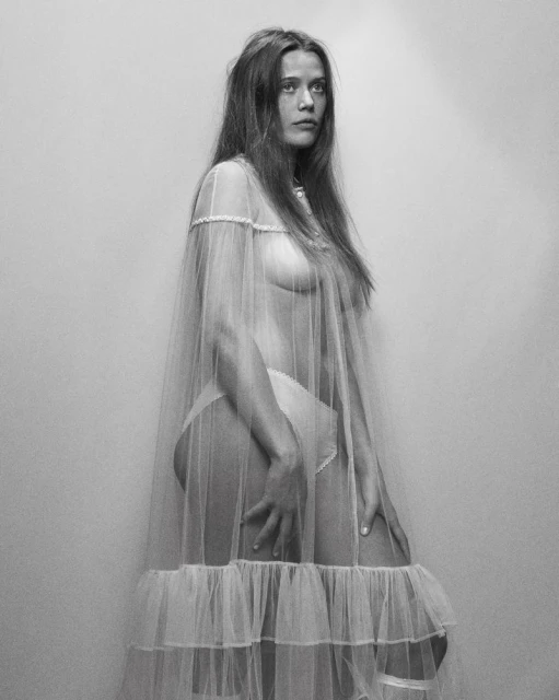 Monochrome portrait of a woman with long hair, partially nude, draped in sheer white fabric against a plain off-white backdrop.