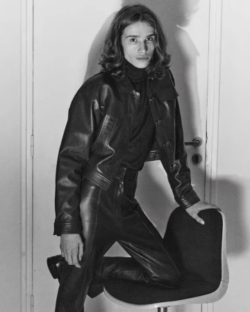 Model Baptiste Vivo in a black leather jacket and leather trousers resting a knee on a swivel chair in a black and white editorial.