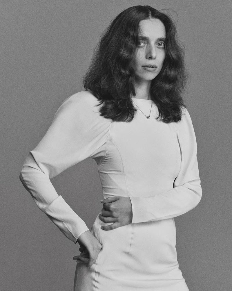 Monochrome portrait of a young woman in a long-sleeved dress against a neutral background, gazing directly at the camera.