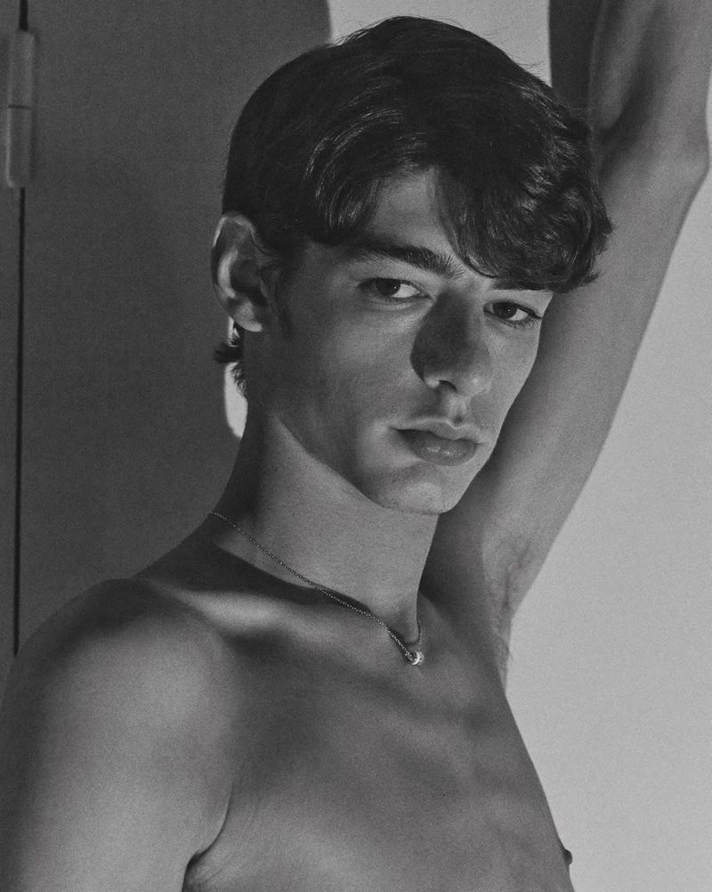 Black and white portrait of shirtless model Baptiste Vivo with an arm raised, wearing a delicate silver chain necklace.