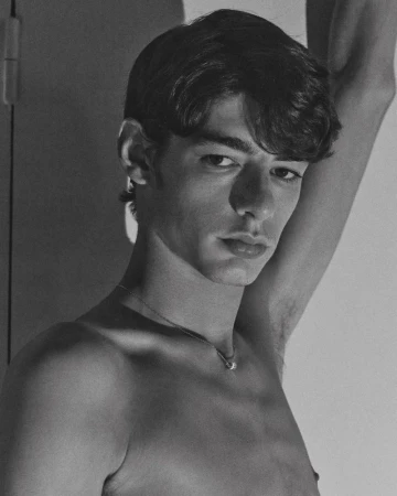 Black and white portrait of shirtless model Baptiste Vivo with an arm raised, wearing a delicate silver chain necklace.