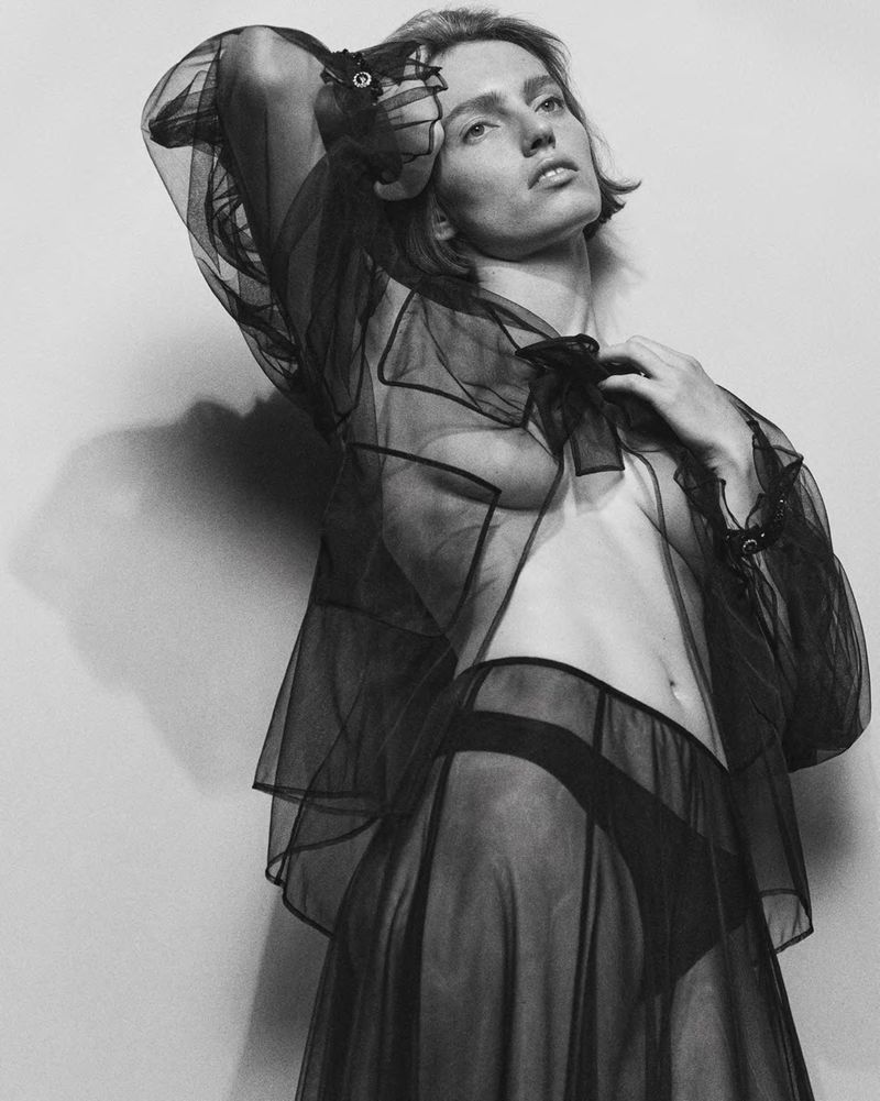 Black and white studio shot of a woman posing in sheer black lingerie, with a neutral facial expression against a plain backdrop.