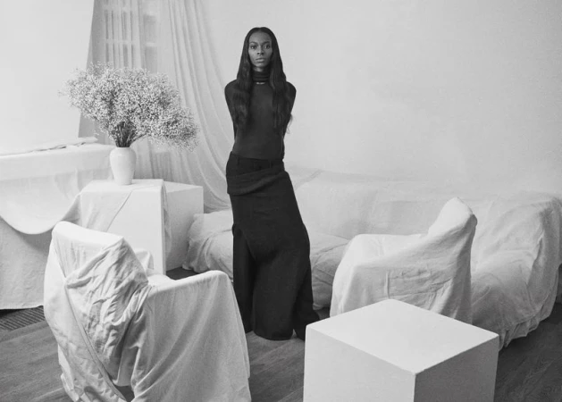 Monochrome fashion portrait of a woman standing amidst white sheet-covered furniture and pedestals with a vase of flowers.