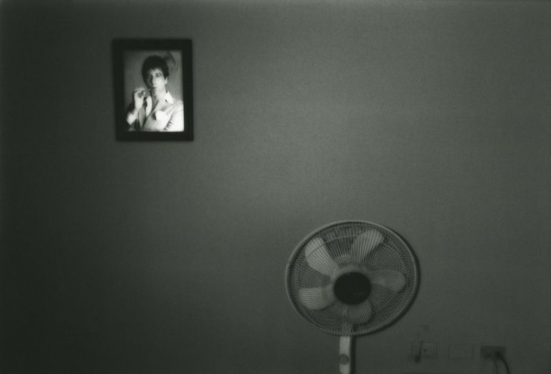Monochrome photograph of an electric fan with a framed portrait of an adult on the wall, creating a vintage, intimate indoor scene.