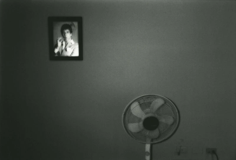 Monochrome photograph of an electric fan with a framed portrait of an adult on the wall, creating a vintage, intimate indoor scene.