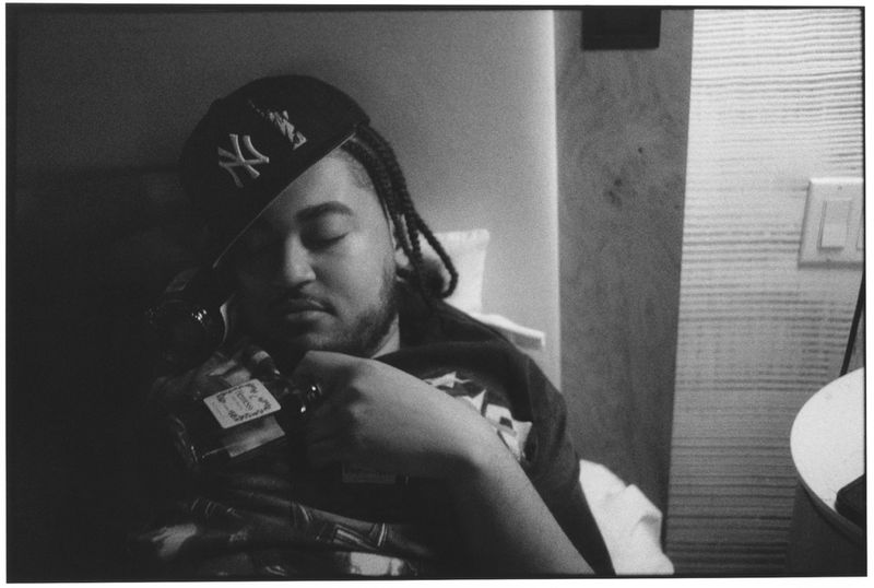 Black and white portrait of Cktrl wearing a New York Yankees baseball cap and holding a bottle while resting against a pillow.