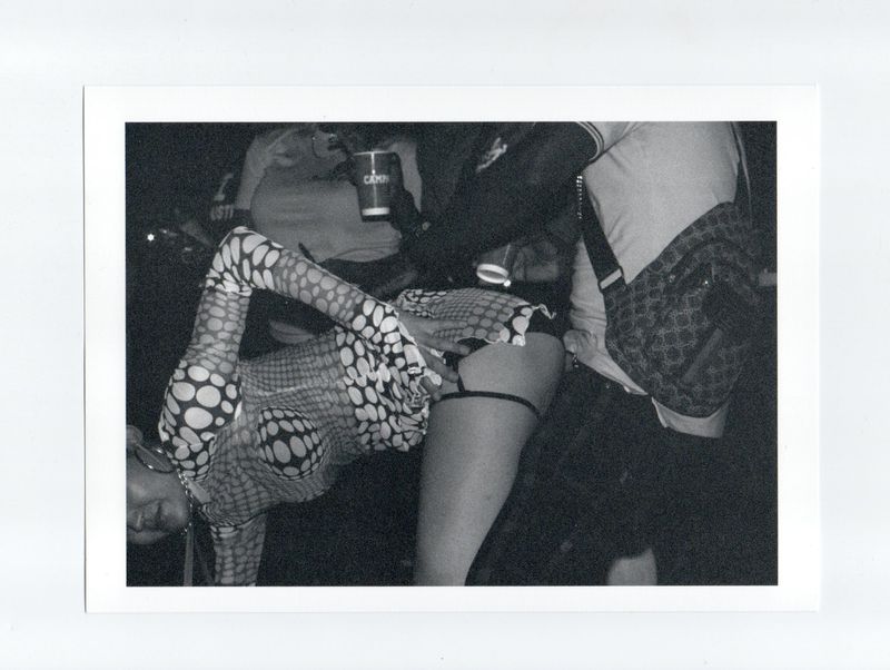 Black and white grainy photo of a woman in a patterned top bending over, partially visible amongst a group of people at a party.