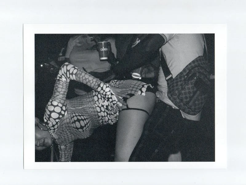 Black and white grainy photo of a woman in a patterned top bending over, partially visible amongst a group of people at a party.